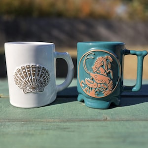 May include: Two ceramic mugs. One is white with a seashell design. The other is teal with a lobster and wheat design. Both mugs have large handles and are on a green surface.