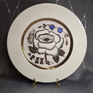 May include: Cream-colored decorative plate featuring a large, stylized white and gray flower with gold accents. The flower is surrounded by gold trim and blue floral accents. The plate is displayed on a gold stand.