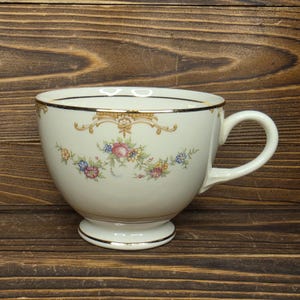 May include: Cream-colored teacup with a floral design featuring pink, blue, and yellow flowers. The cup has a gold rim and a decorative gold accent near the top. The teacup sits on a small, round foot.