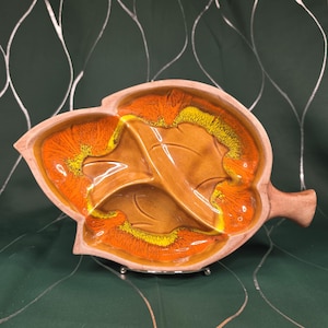 May include: A leaf-shaped serving dish with a handle, featuring a brown base and a glossy interior with orange and yellow accents. The dish is divided into three sections and rests on a silver metal stand.