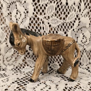 May include: A ceramic donkey figurine in shades of brown, tan, and black. The donkey has a basket on its back and the word "CLOVELLY" is written on its side. The figurine is standing on four legs.