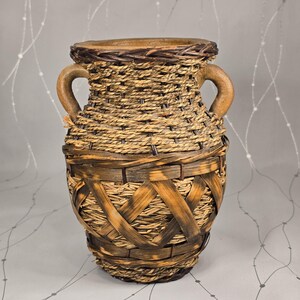 May include: A decorative vase with a woven design. The vase features a light brown woven body with dark brown accents and two curved handles. The top rim is a darker brown, and the base has a woven pattern.