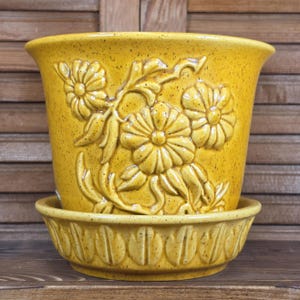 May include: A mustard yellow ceramic plant pot with a speckled finish and a matching saucer. The pot features a raised relief design of three large flowers with leaves, while the saucer has a repeating vertical petal pattern around the exterior.