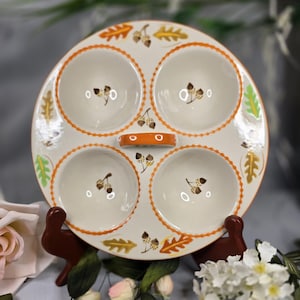 May include: A round, off-white ceramic serving dish with four circular compartments. The dish features an orange handle and is decorated with orange trim, acorn and leaf motifs. The dish is displayed on a brown stand, with flowers in the foreground.