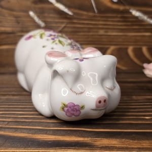 May include: A white ceramic pig figurine with closed eyes, adorned with pink bows and floral accents. The pig has a pink snout and is decorated with purple and pink flowers. The figurine is resting on a wooden surface.
