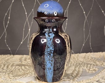 May include: A decorative ceramic vase with a matching lid. The vase is primarily dark purple with a vertical blue stripe and cream-colored accents. The lid is blue and brown. The vase sits on a cream-colored lace surface.
