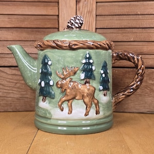 May include: A ceramic teapot with a green base and lid, featuring a brown moose and evergreen trees. The handle and rim are designed to look like tree branches. A pinecone tops the lid. The teapot is a decorative item.