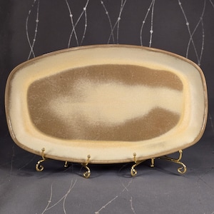 May include: A rectangular ceramic serving platter with rounded corners. The platter has a cream-colored border and a central area with a gradient of light brown to cream. It is displayed on a gold-colored metal stand.
