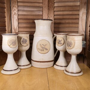 May include: A cream-colored ceramic pitcher and four matching goblets. The pitcher and goblets have a brown rim and base. Each goblet has a leaf design. The pitcher has a circular design with a plant.