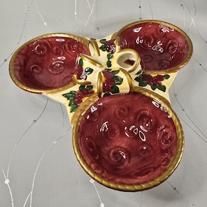 May include: A decorative, tri-sectioned serving dish with a bow-shaped handle. The dish features three burgundy-colored bowls with a gold rim. The bow and base are cream-colored with floral accents. A unique piece for serving snacks or appetizers.
