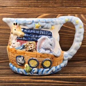 May include: A ceramic pitcher depicting Noah's Ark, featuring a giraffe, elephant, and other animals. The pitcher has a blue handle and is decorated with clouds and stars. The ark is brown with blue accents.