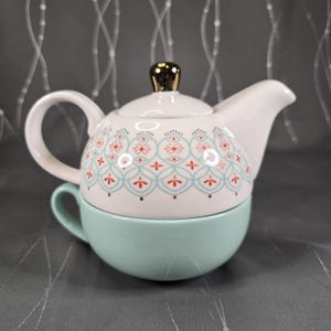 May include: A white ceramic teapot with a gold-colored knob on the lid, decorated with a blue and red floral pattern, sits atop a matching light green teacup. The teapot has a curved handle and spout.
