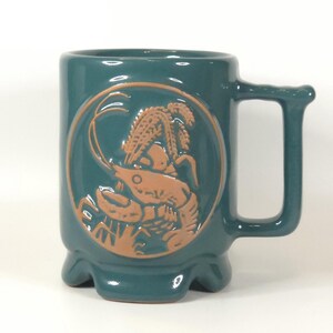 May include: A teal ceramic mug featuring a raised, circular relief design in a light brown tone. The relief depicts a crawfish surrounded by wheat stalks. The mug has a sturdy handle and a base with four small, rounded feet for stability.