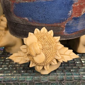 May include: A cream-colored carved ring featuring a sunflower design with a bee resting on the center petals. The sunflower has textured seeds and pointed leaves extending from the sides. The ring is crafted from a solid, matte material.