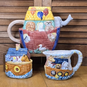 May include: A ceramic Noah's Ark themed tea set. The teapot has an elephant spout and a lid with balloons. The set includes a sugar bowl and a creamer, all decorated with animals and a boat design. The colors are bright and whimsical.