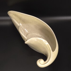 May include: A cream-colored ceramic bowl with a long, tapered, leaf-like shape. The vessel features a smooth, glossy finish with subtle speckled detailing throughout the surface and a distinct curled, scroll-like handle at one end.