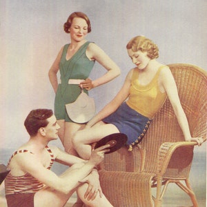 May include: Three people are posing for a photo. A woman in a green swimsuit is holding a white sun hat. Another woman in a yellow top and blue skirt is sitting in a wicker chair. A man in a striped swimsuit is sitting on the floor. The photo is from April 1933.