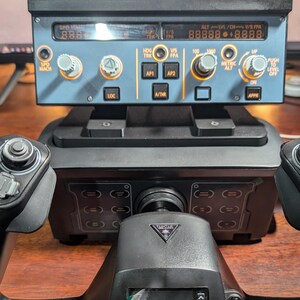 May include: A black flight simulator yoke with a small screen and buttons. The yoke is connected to a black base with a control panel that has a digital display with the text "ALTV/OHX/S FRA 888888888" and "SPO MM 8.8.8".