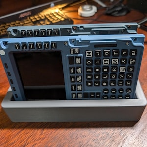 May include: Two blue and gray electronic devices with a keyboard and a screen. The devices are in a gray stand. The keyboard has keys labeled with letters, numbers, and symbols. The screen is black and blank. The devices are on a wooden surface.