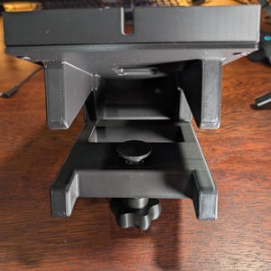 May include: A black, 3D-printed camera mount with a rectangular base and adjustable clamp. The mount features a central screw for securing a camera and a textured surface for grip. The device is designed to attach to a tripod or other support.
