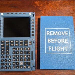 Może przedstawiać: Niebieski i czarny panel sterowania kokpitu samolotu z ekranem i klawiaturą. Panel ma etykietę z napisem "REMOVE BEFORE FLIGHT".