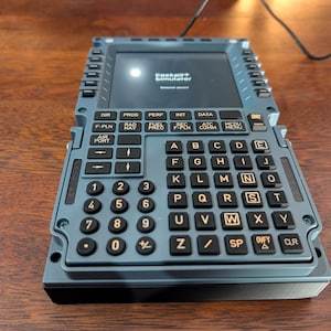 May include: A gray cockpit simulator with a screen displaying text and a keyboard with letters and numbers. The simulator has various function buttons labeled with abbreviations. A black cable is connected to the device.
