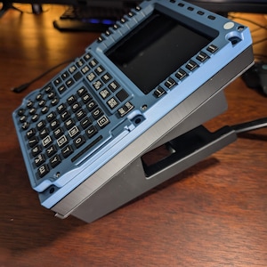 May include: A blue and black electronic device with a keyboard and screen. The device has a rectangular shape with a keyboard featuring letters and numbers. The screen is black. The device is on a black stand.