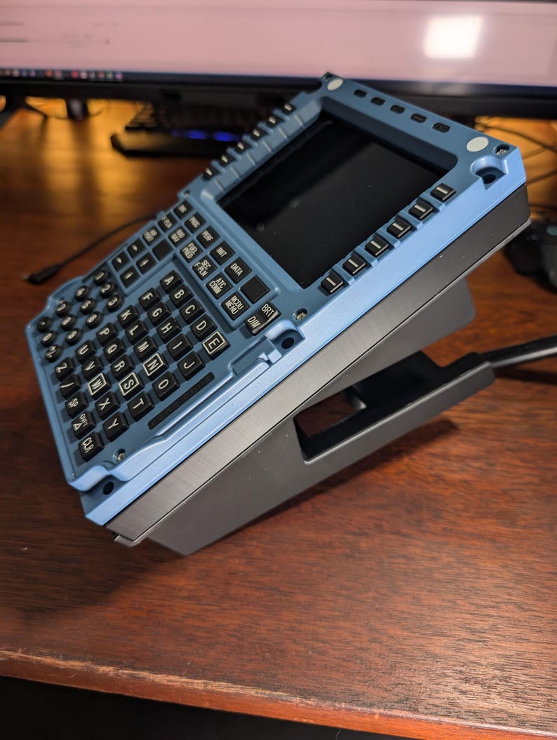 May include: A blue and black electronic device with a keyboard and screen. The device has a rectangular shape with a keyboard featuring letters and numbers. The screen is black. The device is on a black stand.