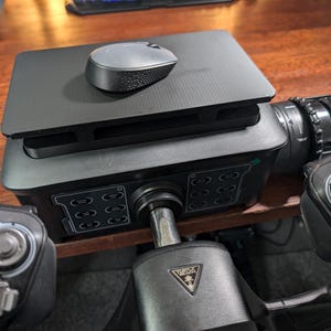 Op de afbeelding: Een zwart gaming stuurwiel met een zwart muismatje en een zwarte muis erop. Het stuurwiel heeft een zwart en groen bedieningspaneel met knoppen en een zwarte en zilveren versnellingspook. Het stuurwiel staat op een houten oppervlak.