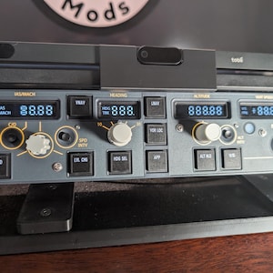 May include: A grey and black flight simulator panel with digital displays showing numbers. The panel features buttons and knobs labelled with aviation terms. A black Tobii device is mounted above the panel. The word "Mods" is visible in the background.