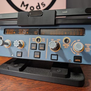 Könnte beinhalten: Ein schwarzes und graues Flugsimulator-Bedienfeld mit mehreren Knöpfen, Tasten und digitalen Anzeigen. Das Panel zeigt Texte wie "SPD MACH", "HDG TRK LAT" und "V/S FPA". Über dem Panel ist eine schwarze Tobii-Sensorleiste montiert. Es steht auf einem schwarzen Ständer.