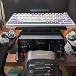 May include: A black Moza flight control yoke featuring a central display with flight checklist text including Before Takeoff and After Takeoff sections. Behind the yoke sits a black Moza control panel and a white mechanical keyboard with purple keys.