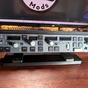 以下が含まれることがあります： グレーと黒の航空機のコックピットパネル。さまざまなボタン、スイッチ、デジタルディスプレイがあります。パネルは黒いスタンドに取り付けられており、背景にはコンピューターモニターとキーボードがあります。「Mods」という単語がモニターに表示されています。