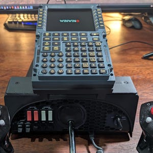 Puede incluir: Un panel de control de simulador de vuelo negro y gris con un teclado azul y amarillo, una palanca de mando negra y dos botones de control negros y rojos. El teclado tiene el texto "WINWINO" en la pantalla.