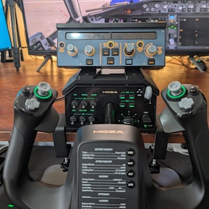 May include: A black flight simulator control panel with multiple buttons, knobs, and a display screen. The panel is labeled "MOZA" and includes a yoke, throttle, and other flight controls. The background shows a computer screen displaying a flight simulation.
