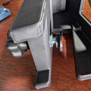 May include: A black 3D-printed object with a rectangular frame and a support structure. A bolt and nut secure a component to the frame. The object is on a wooden surface.