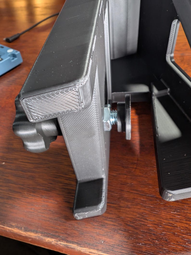 May include: A black 3D-printed object with a rectangular frame and a support structure. A bolt and nut secure a component to the frame. The object is on a wooden surface.