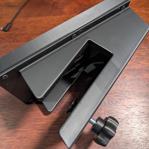 May include: A black, angled computer monitor mount with a textured surface. The mount features a clamp with a black, star-shaped knob for adjusting the angle. The mount is designed to attach to a desk or other surface.
