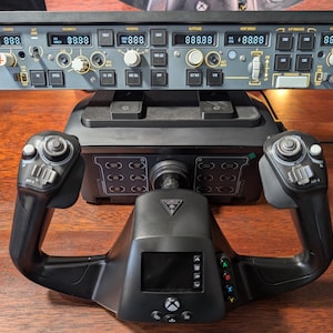 May include: A black flight simulator yoke with a small screen and buttons. The yoke is connected to a black base with a panel of buttons and a throttle lever. The yoke is on a wooden surface. The panel has a digital display with the numbers 88888.