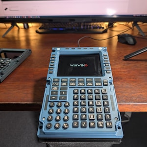 May include: A blue electronic device with a screen displaying "WINWING" and a keyboard with letters and numbers. The device has multiple buttons and is on a wooden surface. Another similar device is visible in the background.