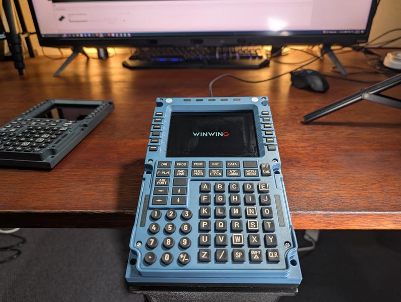 May include: A blue electronic device with a screen displaying "WINWING" and a keyboard with letters and numbers. The device has multiple buttons and is on a wooden surface. Another similar device is visible in the background.
