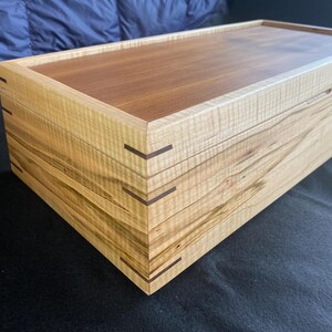 May include: A wooden box with a light brown top and a light brown and tan bottom. The box has a natural wood grain pattern and is made of multiple layers of wood. The box has a dark brown trim around the edges.