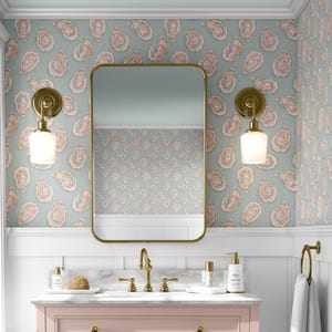May include: A bathroom with a pink vanity, gold fixtures, and a large rectangular mirror. The walls are decorated with wallpaper featuring a pattern of pink and white oyster shells on a light blue background. Two gold sconces flank the mirror.