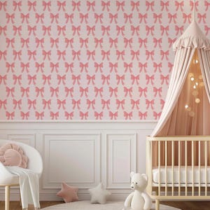 May include: A pink and white nursery with a white crib canopy, a white crib with a white mattress, a white armchair with a pink pillow, and a pink and white patterned wallpaper with pink bows.