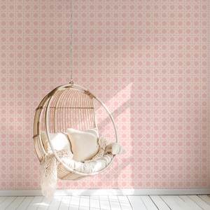 May include: A hanging wicker chair with white cushions and a cream-colored throw blanket. The chair is suspended from a chain against a pink and white patterned wallpaper. The floor is white.