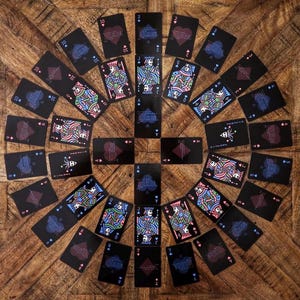 Puede incluir: Un conjunto de cartas de juego dispuestas sobre una superficie de madera. Las cartas, con fondo negro, presentan palos y figuras coloridas. La disposición circular resalta los diferentes símbolos y números, creando un efecto visual atractivo.