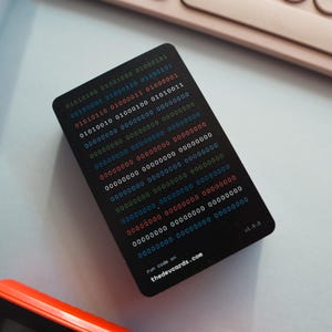 Peut inclure: Une carte noire avec du texte blanc et des chiffres colorés. Le texte indique "run code on thedevcards.com v1.0.0". La carte comporte une série de code binaire imprimé dessus.