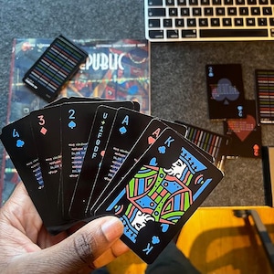 May include: A hand holds a deck of playing cards with black backgrounds and colourful designs. The cards include the numbers 2, 3, and 4, the letter U, and the letter A. The Ace of Spades card has a colourful illustration of a king with a crown.