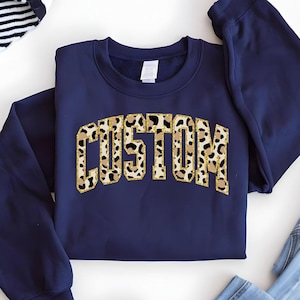 May include: Navy blue sweatshirt with a gold and brown leopard print "CUSTOM" graphic.