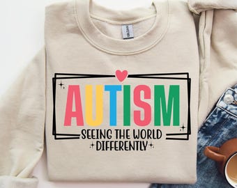 May include: A beige sweatshirt with the text "AUTISM - SEEING THE WORLD DIFFERENTLY" in a black frame. The word "AUTISM" is in rainbow colors.